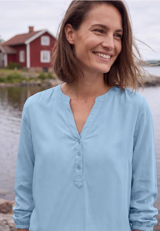 Eine lächelnde Frau in blauer Bluse steht vor einer roten Häuserkulisse am Wasser.