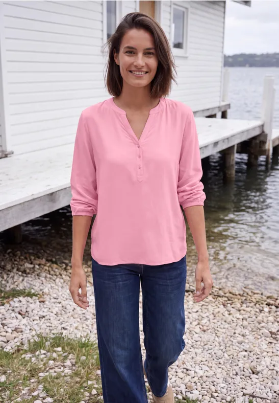 Vrouw in roze blouse en blauwe jeans glimlacht terwijl ze bij een bootshuisje aan het water staat.