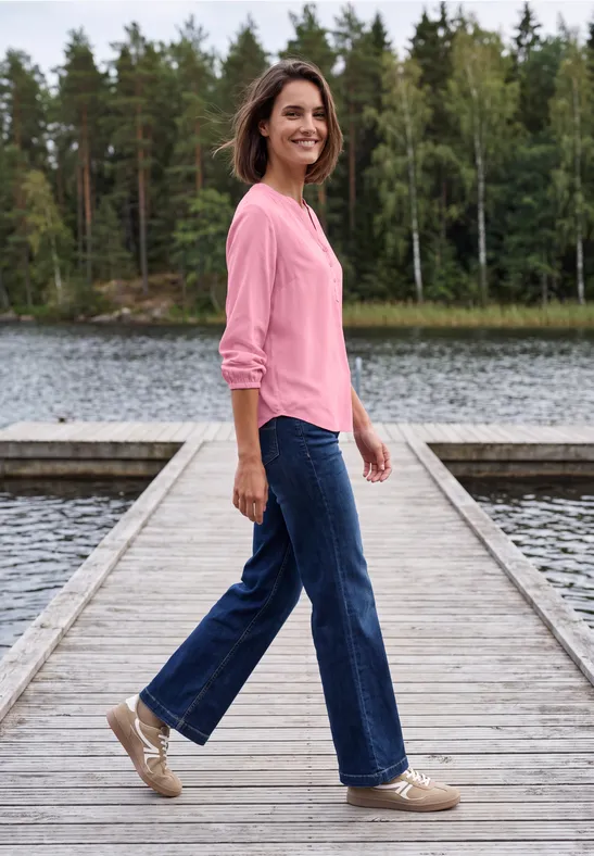 Een vrouw in een roze trui en spijkerbroek wandelt lachend over een houten steiger boven een rustig meer.