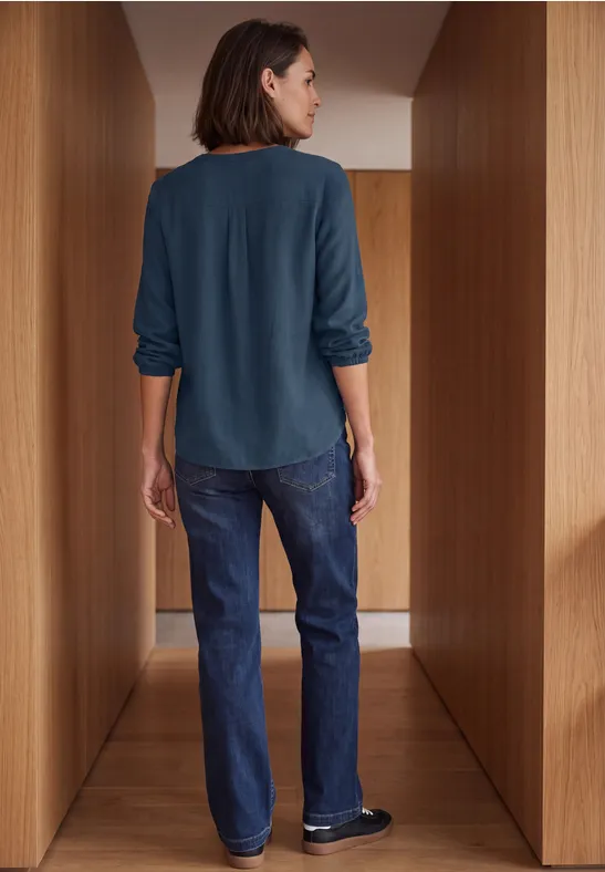 Eine Frau steht in einem hölzernen Flur, von hinten zu sehen, trägt ein dunkelgrünes Oberteil und blaue Jeans.