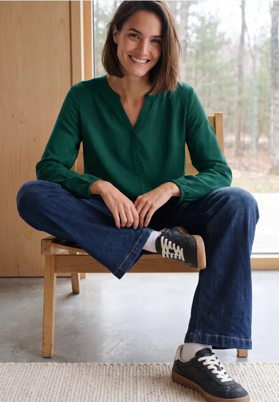 Eine lächelnde Frau sitzt im Schneidersitz auf einem Stuhl, trägt grüne Bluse, Jeans und schwarze Sneaker.