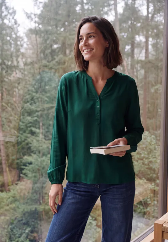 Frau in grüner Bluse steht lächelnd vor einem Waldpanorama, hält ein Buch und einen Stift in der Hand.