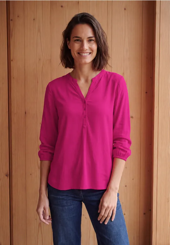 Een glimlachende vrouw in een roze blouse en spijkerbroek voor een houten wand.
