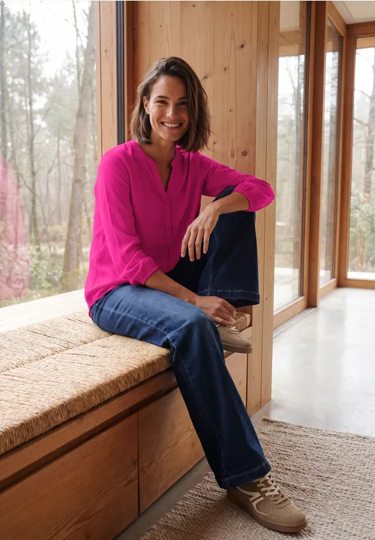 Glimlachende vrouw in roze blouse en spijkerbroek zit ontspannen op een vensterbank in een houten interieur.