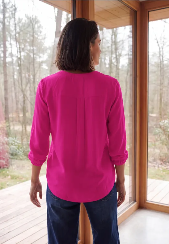 Vrouw van achteren in een felroze blouse en spijkerbroek, voor een glazen deur met boszicht.