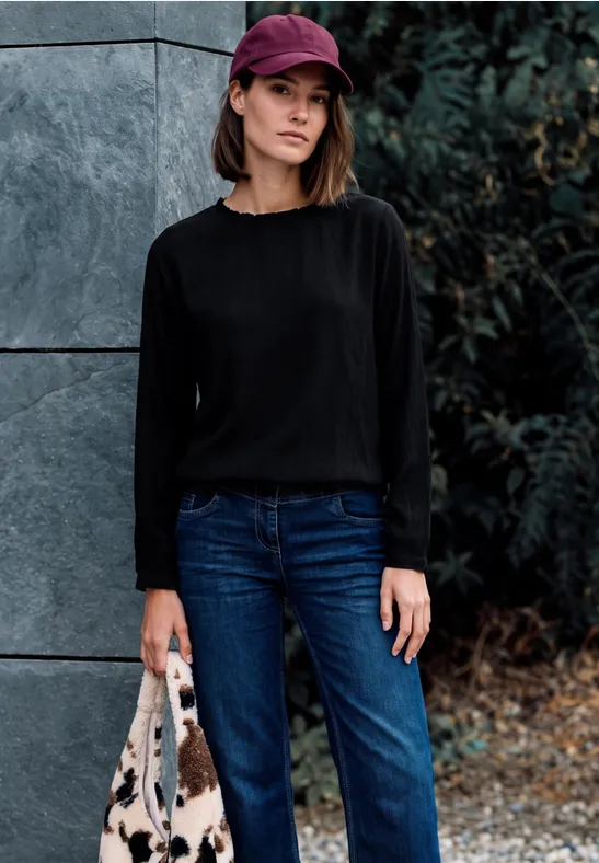 Frau mit schwarzem Pullover, Jeans, roter Mütze und Tasche steht an einer Wand im Freien.