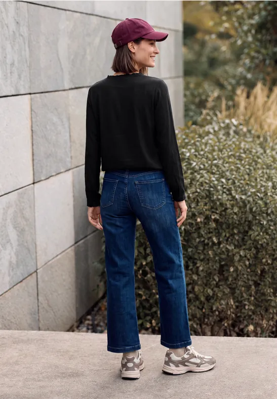 Person trägt schwarzen Pullover, blaue Jeans, graue Sneaker und Kappe, steht seitlich vor Steinmauer und Büschen.