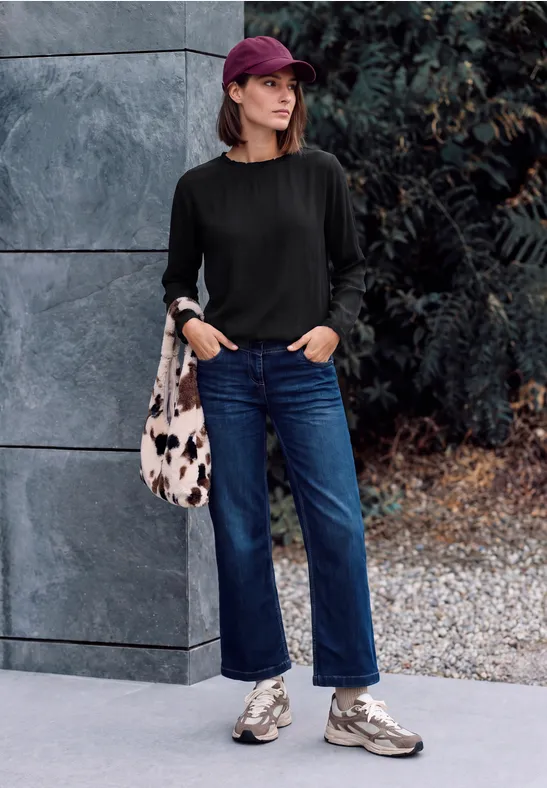 Frau in schwarzem Pullover, blauen Jeans, Sneakers, Baseballcap und Tasche lehnt an einer Fassade.