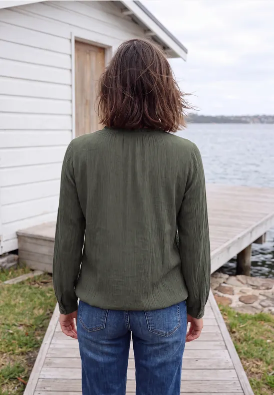 Person in grüner Jacke und Jeans steht auf einem Bohlenweg vor einer weißen Hütte am Wasser.