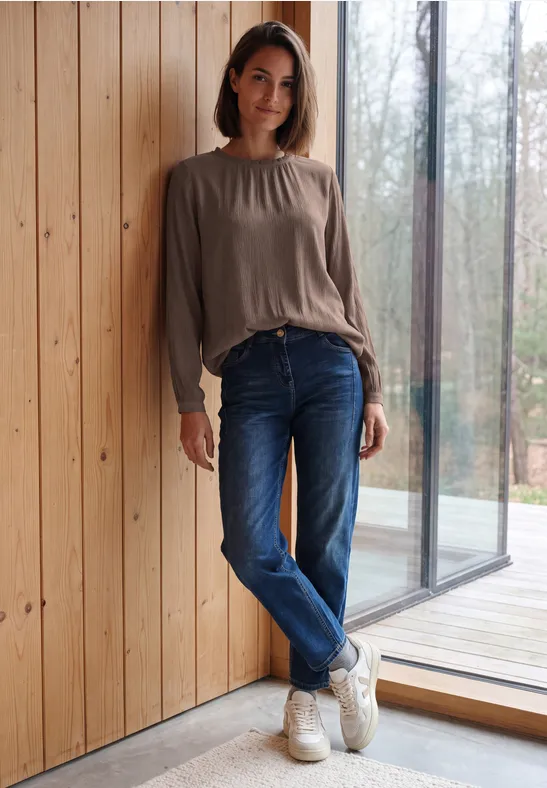 Eine junge Frau lehnt entspannt an einer Holzwand, trägt Jeans, Sneakers und ein lockeres Oberteil.