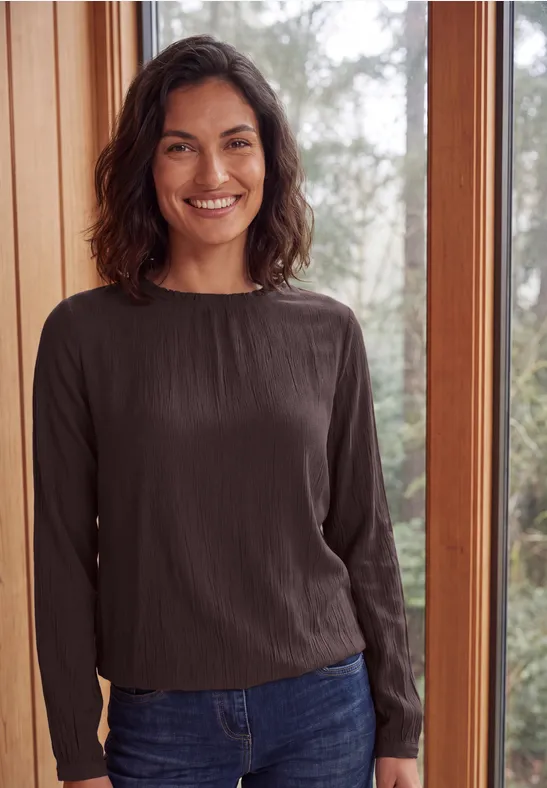 Femme souriante en haut à manches longues et jean, debout devant une grande fenêtre donnant sur la verdure.