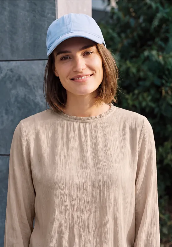 Une femme souriante porte un haut beige et une casquette bleu clair, devant un mur et des arbustes.