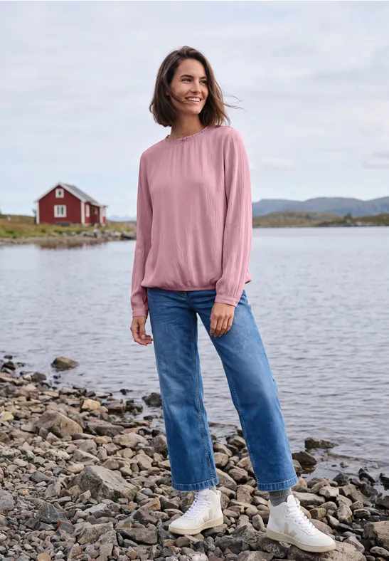 Frau in Jeans und rosa Pullover steht am Ufer vor See und roter Hütte.