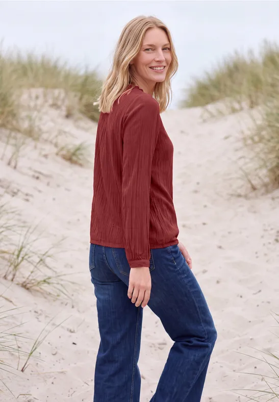 Une femme blonde en pull rouge et jean se tient sur des dunes de sable au bord de la mer et sourit.