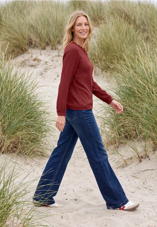 Une femme blonde souriante, en pull rouge et jean bleu, marche sur des dunes de sable avec des herbes.
