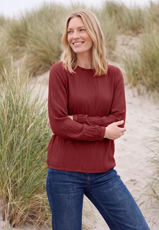 Femme en pull rouge et jean, souriante, debout sur une dune de sable.