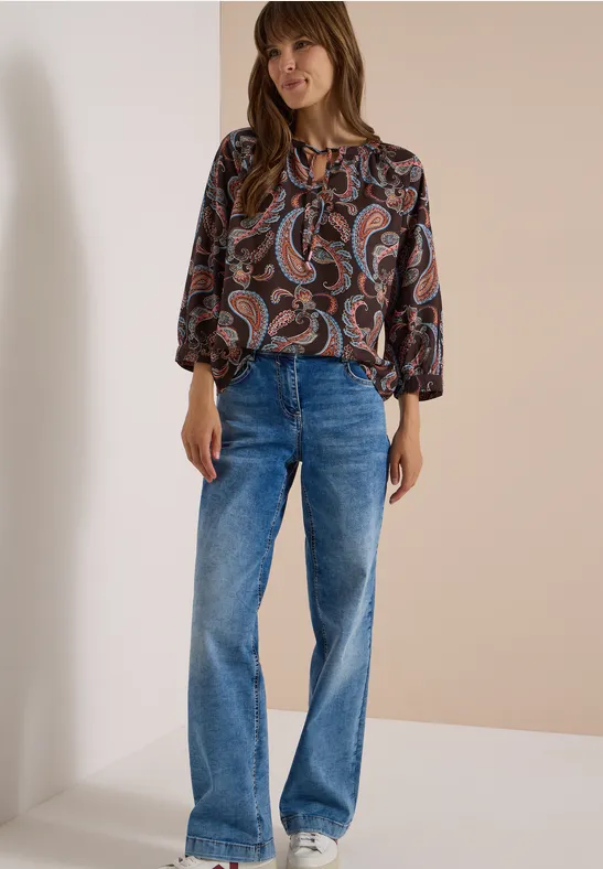 Frau in weicher Paisleybluse und weiten Jeans steht vor cremefarbenem Hintergrund.