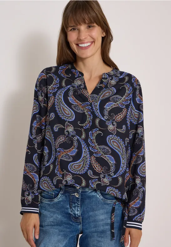 Eine lächelnde Frau in dunkler Paisley-Bluse und Jeans.