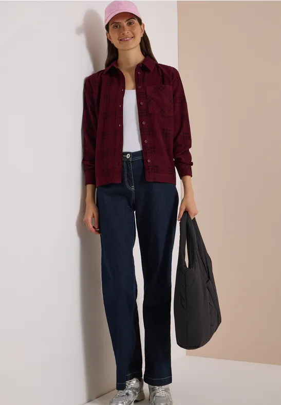 Frau in rotem Karohemd, weißem Top, dunklen Jeans, rosa Cap und schwarzer Tasche vor hellem Hintergrund.
