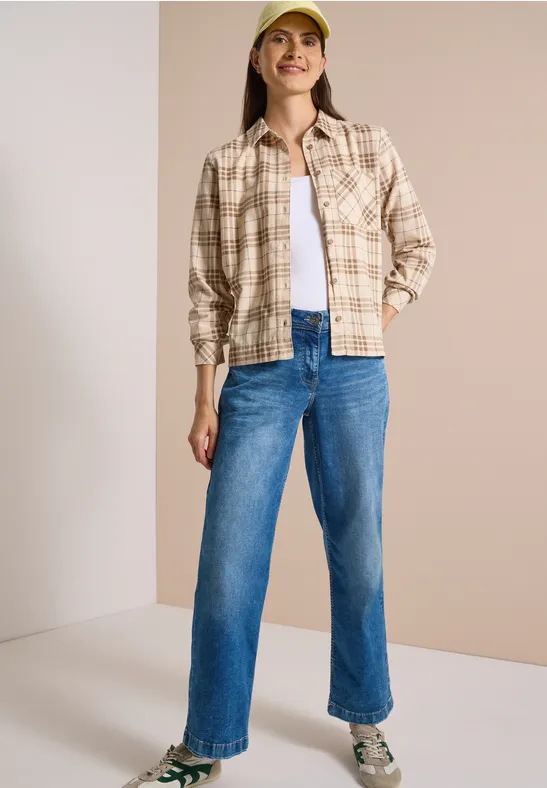 Frau in beiger karierten Hemdjacke, weißem Top, blauen Jeans und gelber Kappe vor neutralem Hintergrund.