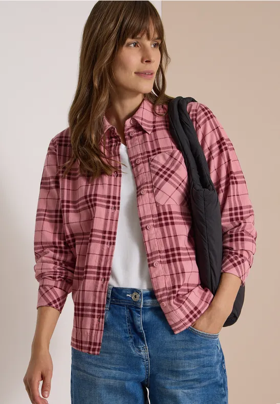 Frau mit pink-karierter Bluse, weißem Top, Jeans und Schultertasche blickt seitlich.