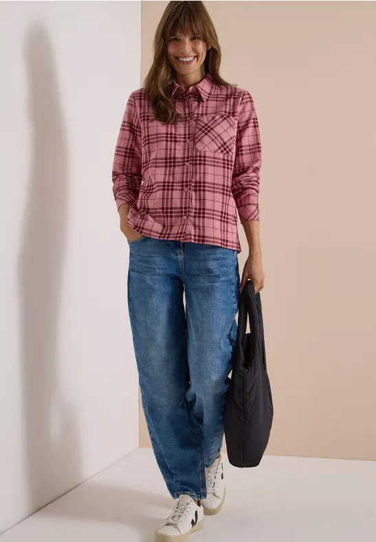 Eine Frau trägt ein pink kariertes Hemd, blaue Jeans und weiße Sneaker, hält eine schwarze Tasche.