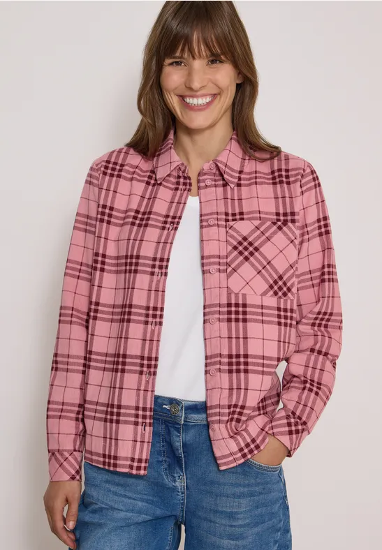 Frau in rosa kariertem Hemd, weißem T-Shirt und Jeans lächelt in die Kamera.