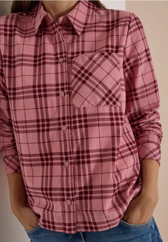 Person trägt pink kariertes Hemd mit Brusttasche, Hände in Jeans-Taschen.