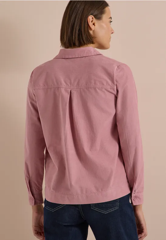 Rückansicht einer Frau mit hellrosa Hemd und Jeans vor beigem Hintergrund.