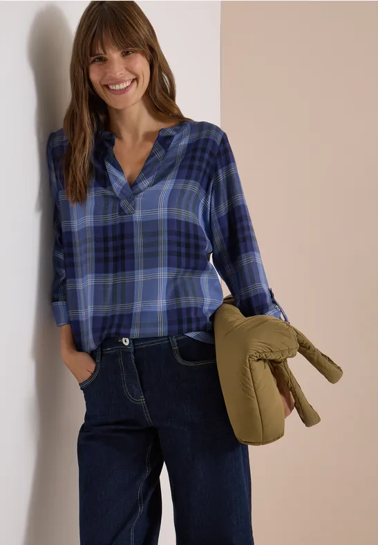 Eine lächelnde Frau in blauer Karo-Bluse und Jeans hält eine grüne gesteppte Jacke.