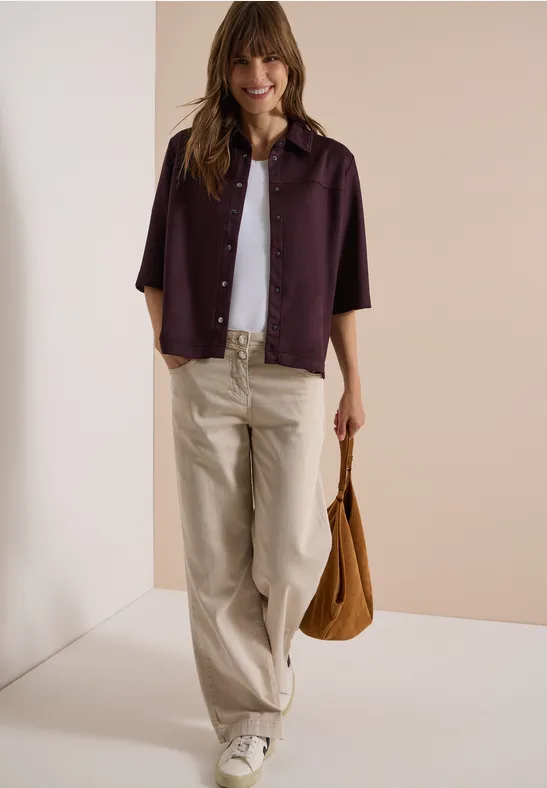 Frau trägt weiße Bluse, lila Hemdjacke, beige Hose und braune Tasche vor neutralem Hintergrund.
