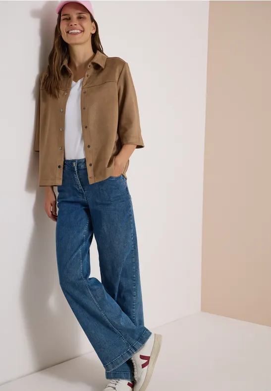 Frau in weiter Jeans, weißem Shirt, brauner Jacke und Sneakern lehnt lächelnd an der Wand.
