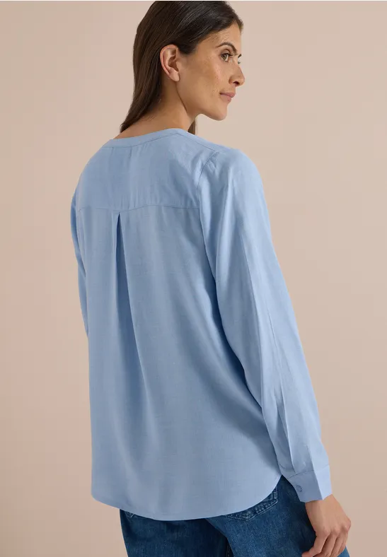 Frau von hinten in hellblauer Bluse und Jeans vor neutralem Hintergrund.