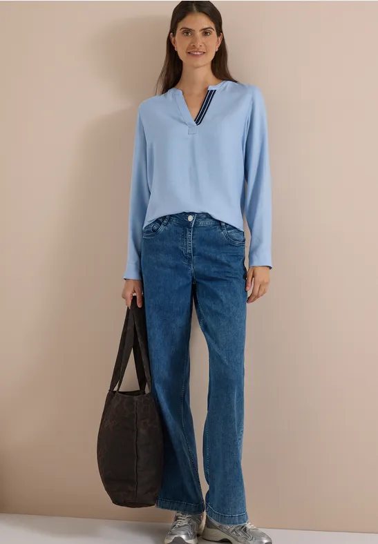 Frau in hellblauer Bluse, blauen Jeans, beigen Sandalen und großer dunkler Tasche vor beigem Hintergrund.