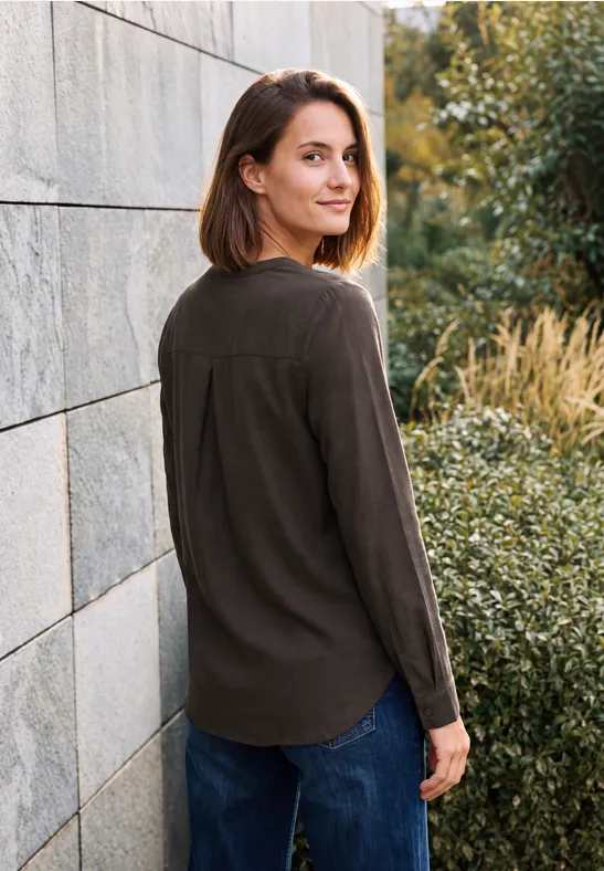 Une femme souriante en tenue décontractée se tient dehors près d'un mur et de plantes, regardant par-dessus son épaule.