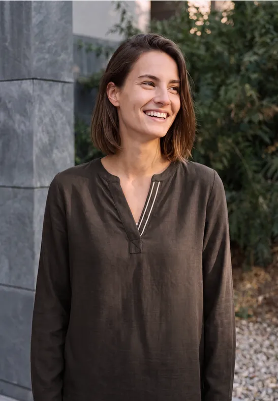 Une femme souriante en chemise marron se tient devant un mur de pierre et des plantes.