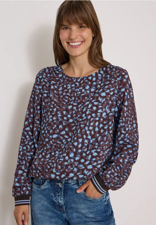 Vrouw lacht in bruin-blauw dessin top en blauwe jeans tegen een lichte achtergrond