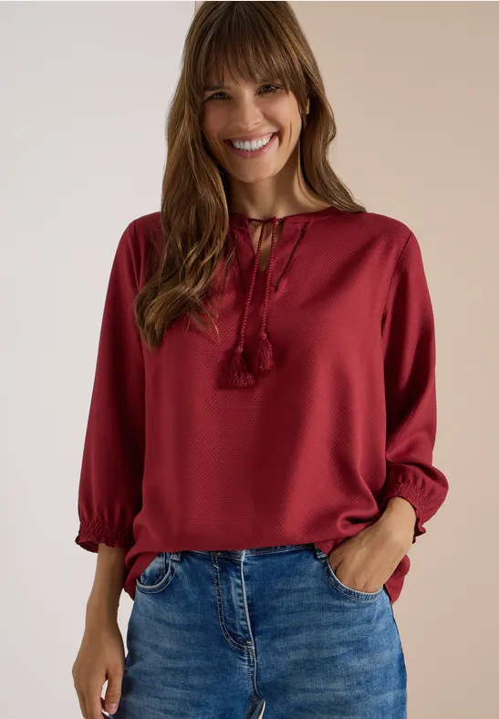 Fröhliche Frau in roter Bluse und Jeans, Hände in Taschen, vor hellem Hintergrund.