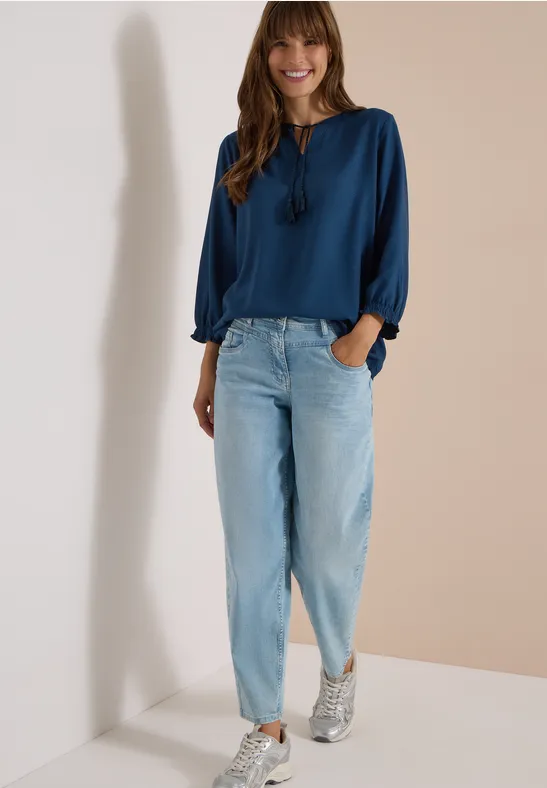 Eine lächelnde Frau lehnt an einer weißen Wand, trägt eine blaue Bluse, helle Jeans und Sneaker.