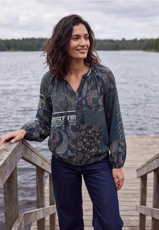 Frau in gemusterter Bluse und Jeans auf hölzernem Steg am See, lächelnd.