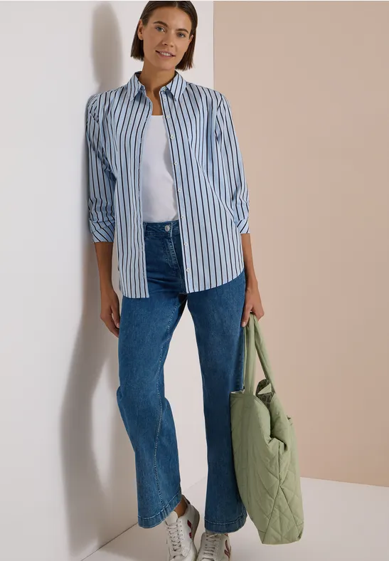 Une femme pose contre un mur, portant une chemise rayée, un débardeur blanc, un jean bleu et un sac vert clair.