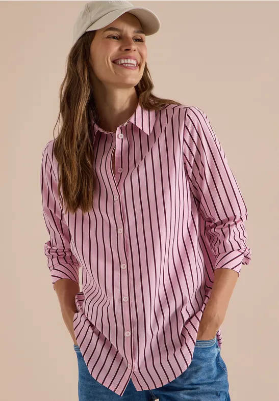 Femme souriante portant une chemise rose à rayures blanches, un jean bleu et une casquette beige clair.