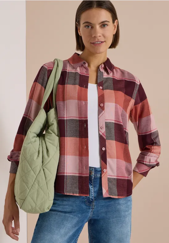 Femme en chemise à carreaux, jean et tee-shirt blanc porte un sac matelassé vert à l'épaule.