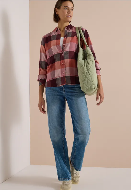 Personne porte chemise à carreaux, jean bleu et sac vert en bandoulière sur fond clair.