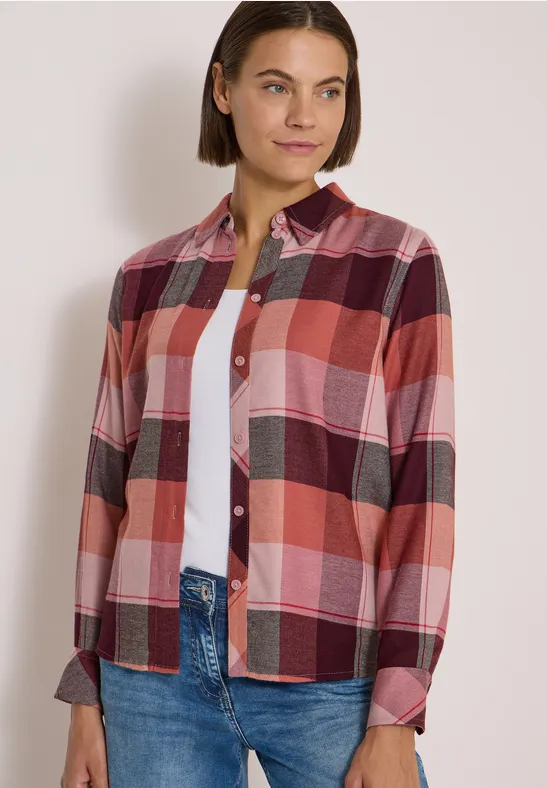 Femme portant une chemise à carreaux rose et rouge sur un haut blanc et un jean, souriant légèrement.