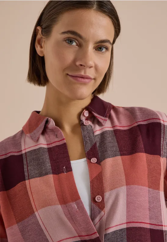 Femme aux cheveux bruns courts porte une chemise à carreaux rouges et regarde la caméra avec un léger sourire.