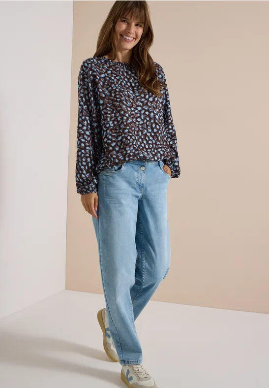 Een lachende vrouw in een kleurrijk blouse en lichte spijkerbroek staat nonchalant voor een neutrale achtergrond.
