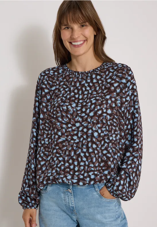 Jonge vrouw met een gebloemde blouse en jeans, glimlachend voor een lichte achtergrond.