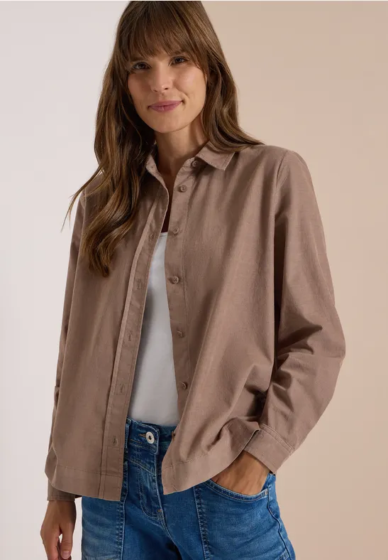 Femme portant une chemise marron, un débardeur blanc et un jean, les mains dans les poches, souriante.