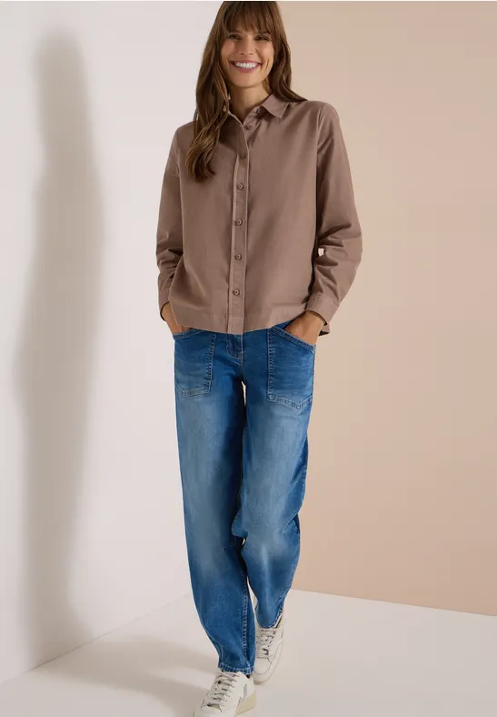 Une femme souriante portant une chemise marron et un jean bleu devant un mur beige.